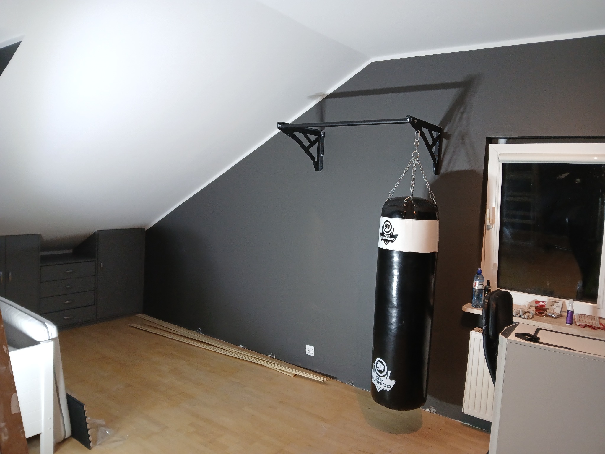 Szary pokój na poddaszu z workiem bokserskim na czarnym drążku, drewnianą podłogą i szarą ścianą. Widoczne listwy przypodłogowe i szafki w tle.