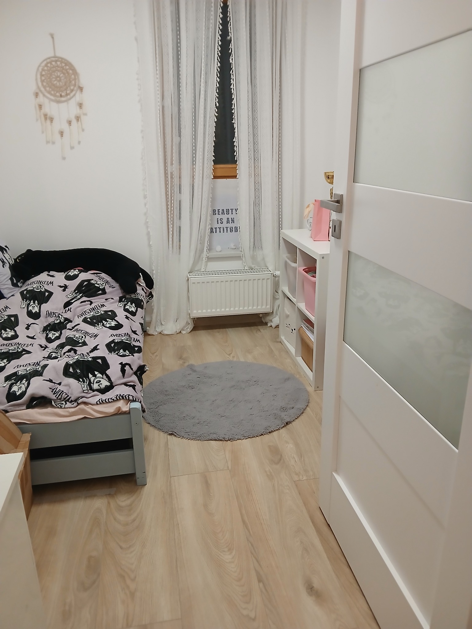 Pokój dziecięcy z łóżkiem w stylu Wednesday, okrągłym dywanem i drewnianą podłogą. Widoczne białe drzwi i dekoracje na ścianie.