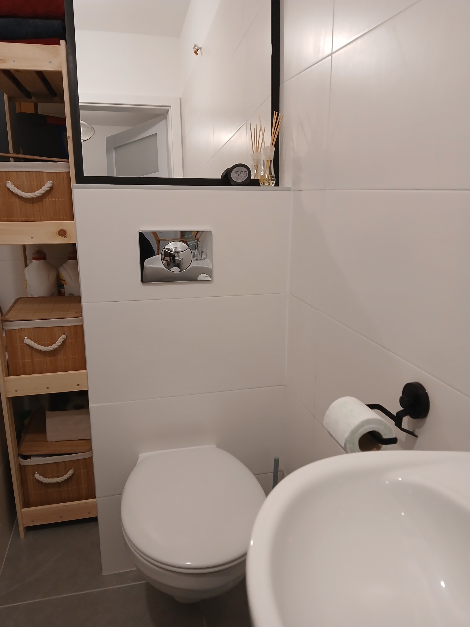 Nowoczesna łazienka z białymi płytkami, wiszącą toaletą i umywalką. Widoczne bambusowe półki i czarne akcenty, takie jak uchwyt na papier toaletowy i rama lustra.