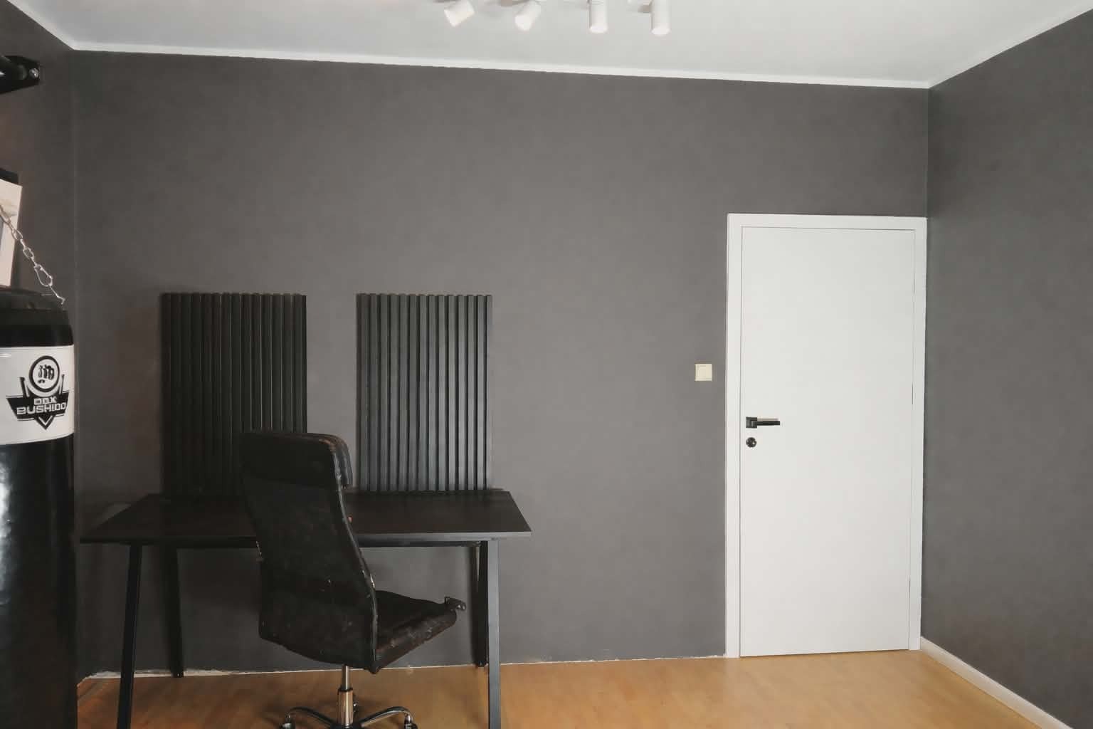 Szara ściana po szpachlowaniu z białymi drzwiami, czarnym biurkiem i workiem bokserskim. Minimalistyczne wnętrze po remoncie, widoczne panele dekoracyjne.