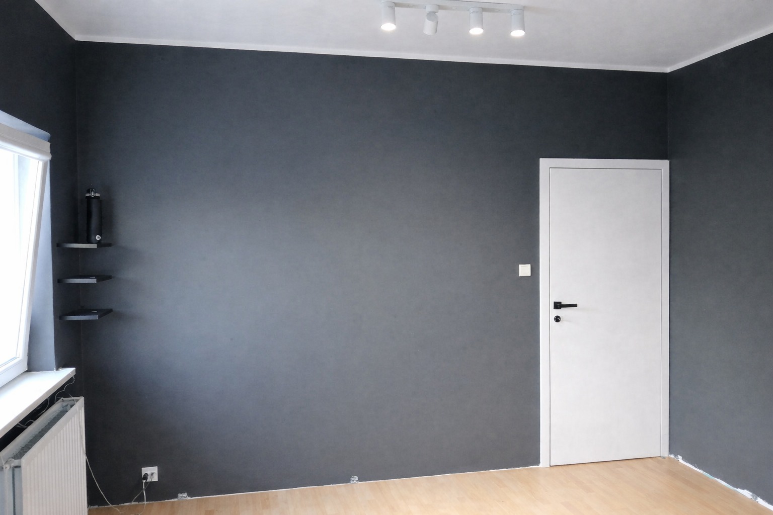 Pokój po remoncie: szare ściany, jasna podłoga, białe drzwi i listwy. Na ścianie półki i lampa sufitowa. Widoczne gniazdko elektryczne i grzejnik.
