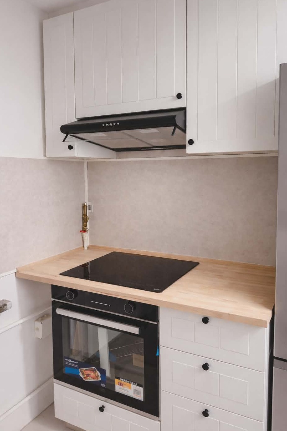 Biała zabudowa kuchenna z drewnianym blatem, czarną płytą indukcyjną i piekarnikiem. Nad blatem szafki i okap. Widoczny fragment instalacji gazowej.