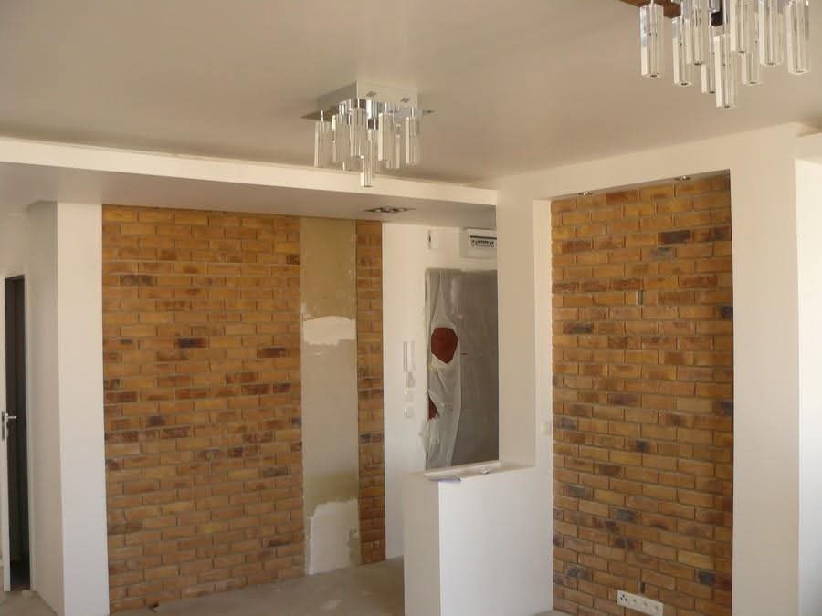 Wykończenie wnętrza z dekoracyjną ścianą z cegły klinkierowej, oświetloną nowoczesnymi lampami. Fragment ściany przygotowany do szpachlowania. Widoczne gniazdka elektryczne.
