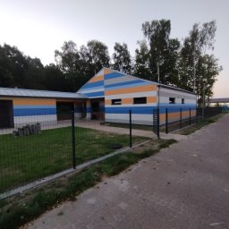 MULTIDOM EXPERT SPÓŁKA Z OGRANICZONĄ ODPOWIEDZIALNOŚCIĄ - Nowo pomalowany budynek z poziomymi pasami w kolorach niebieskim, pomarańczowym, szarym i białym, otoczony zielonym trawnikiem i czarnym ogrodzeniem z siatki, widoczny z chodnika z kostki brukowej.