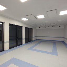 MULTIDOM EXPERT SPÓŁKA Z OGRANICZONĄ ODPOWIEDZIALNOŚCIĄ - Pusta sala z wykończonymi ścianami w odcieniach bieli i szarości, z dużymi oknami z ciemnymi ramami, podwieszanym sufitem z panelami akustycznymi i podłogą z wzorem w niebieskie prostokąty.