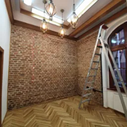 Pokój po remoncie z ceglaną ścianą, parkietem w jodełkę, dekoracyjnymi lampami i rozstawioną drabiną aluminiową.
