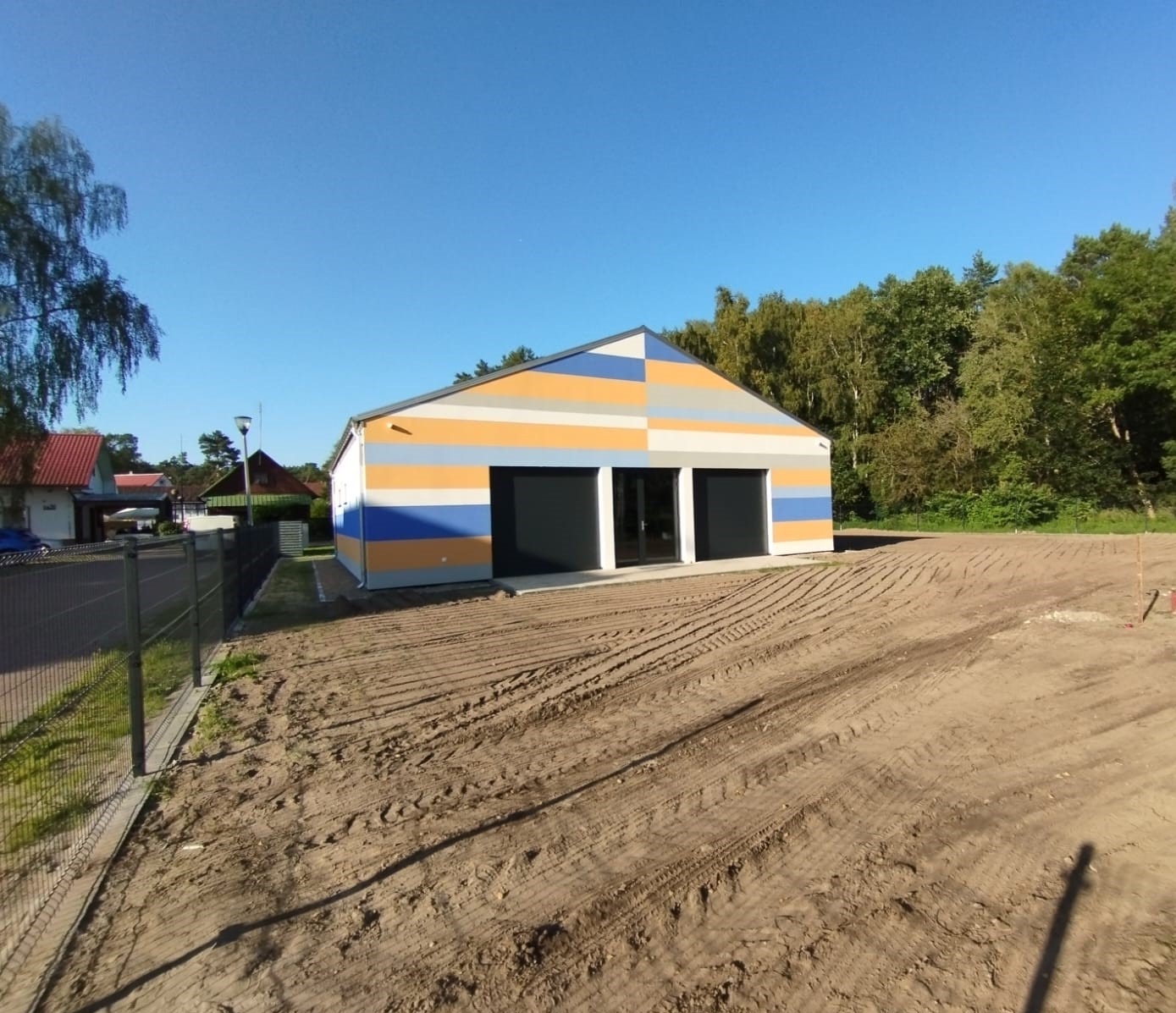 Nowo wybudowany budynek w Szczecinie z pomalowaną w poziome pasy fasadą w kolorach żółtym, pomarańczowym i niebieskim. Teren wokół budynku wyrównany świeżo spulchnioną ziemią.