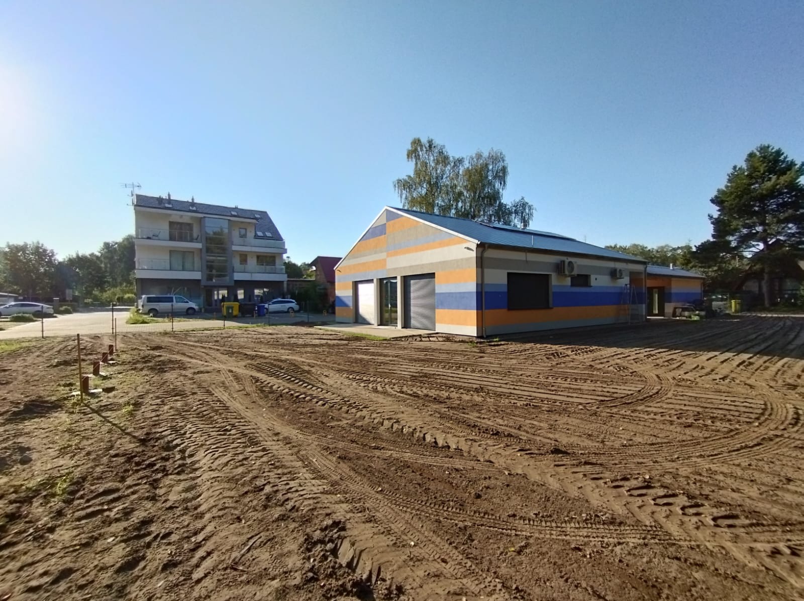 Nowo wybudowany budynek z garażem, pomalowany w poziome pasy koloru pomarańczowego, szarego i niebieskiego, na działce z przygotowaną ziemią pod trawnik.