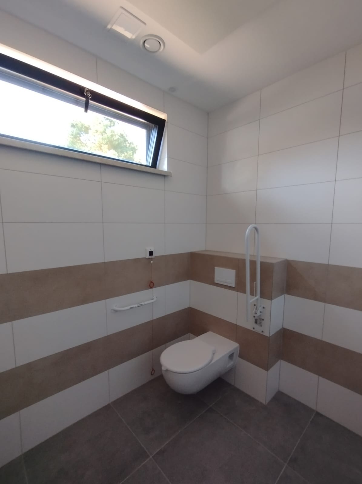 Nowoczesna łazienka z podwieszaną toaletą, uchwytami dla niepełnosprawnych i poziomymi pasami płytek w kolorze białym i beżowym, oświetlona naturalnym światłem z okna.