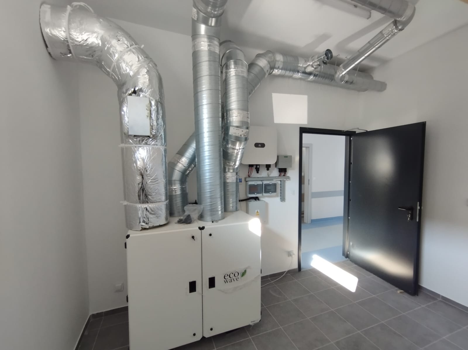 Wnętrze pomieszczenia z zamontowaną centralą wentylacyjną Eco Wave, widoczne srebrne rury wentylacyjne pod sufitem oraz czarne drzwi wejściowe.