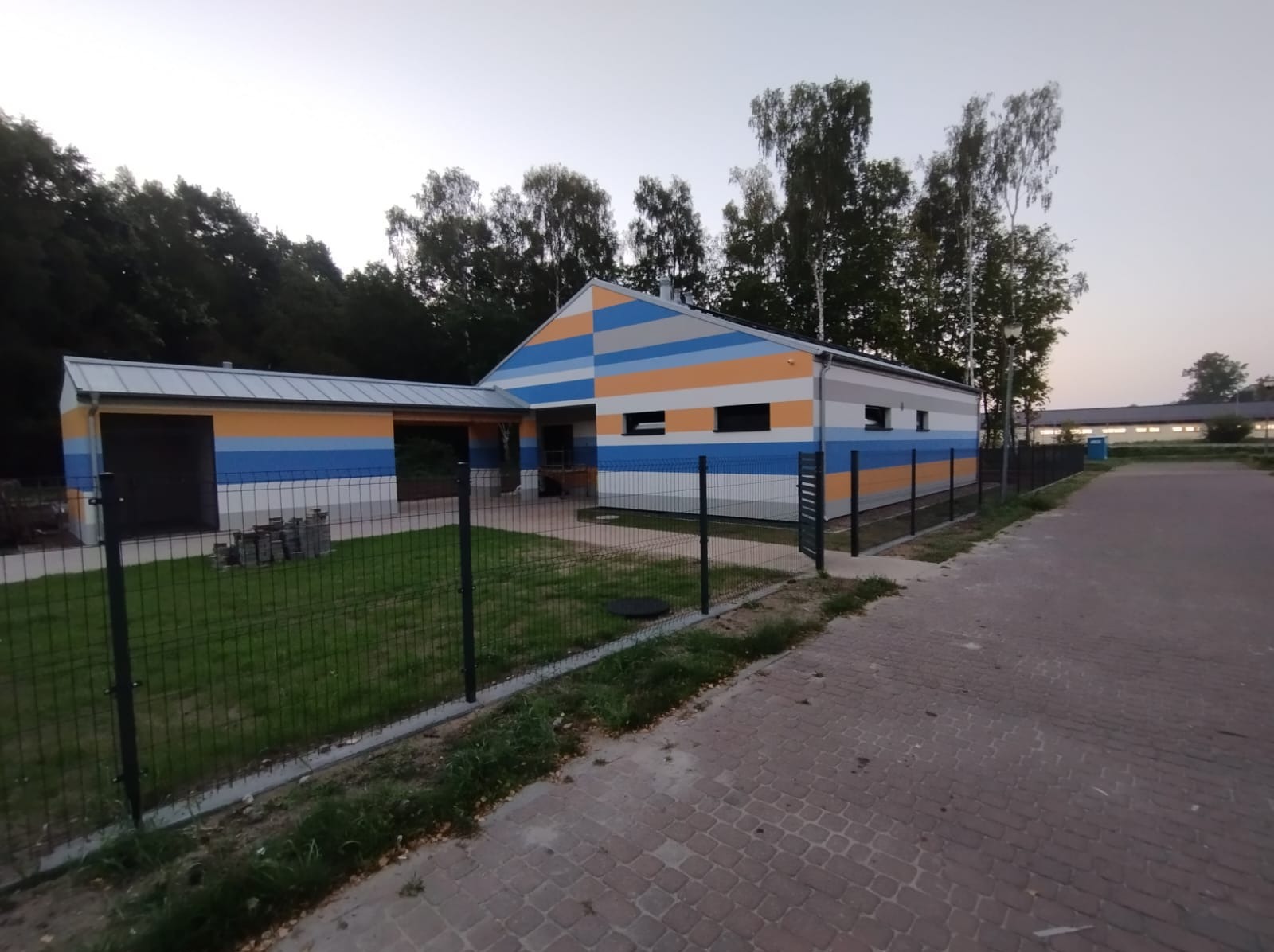 Nowo pomalowany budynek z poziomymi pasami w kolorach niebieskim, pomarańczowym, szarym i białym, otoczony zielonym trawnikiem i czarnym ogrodzeniem z siatki, widoczny z chodnika z kostki brukowej.
