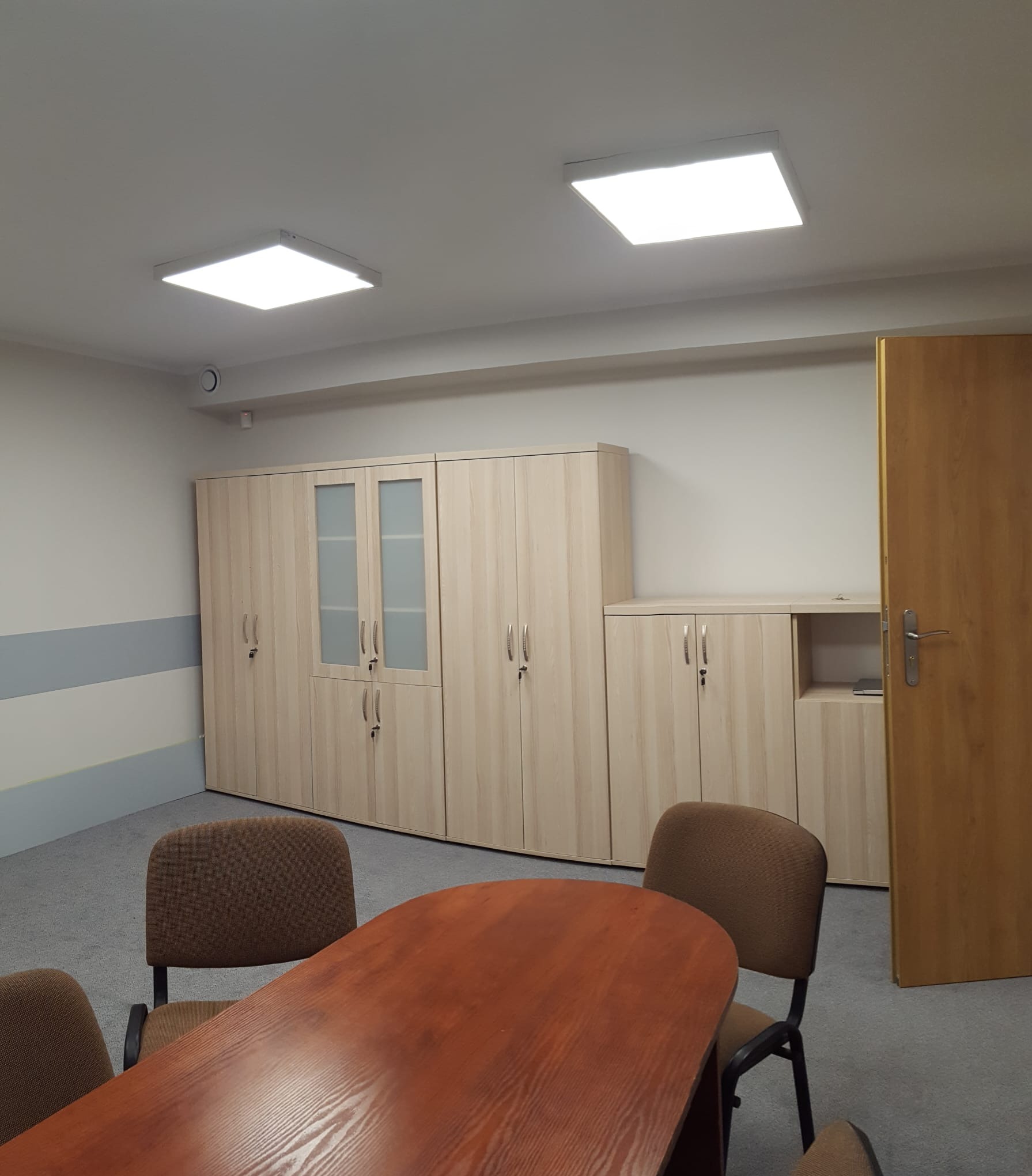 Jasne biuro z drewnianym stołem konferencyjnym, brązowymi krzesłami i szafami z jasnego drewna z metalowymi uchwytami, oświetlone kwadratowymi lampami sufitowymi.