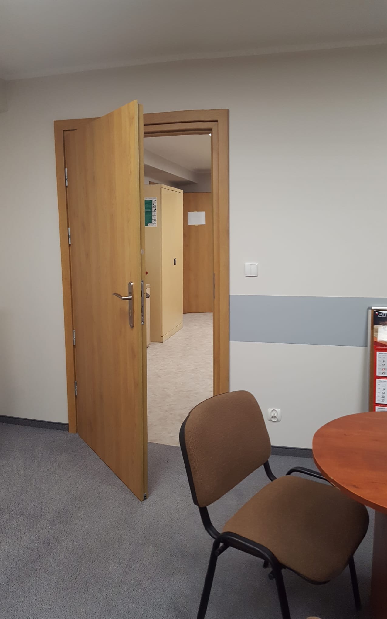 Wnętrze biura z otwartymi drewnianymi drzwiami, widoczne biurko i krzesło na tle szarej wykładziny i białych ścian z szarym pasem ochronnym.