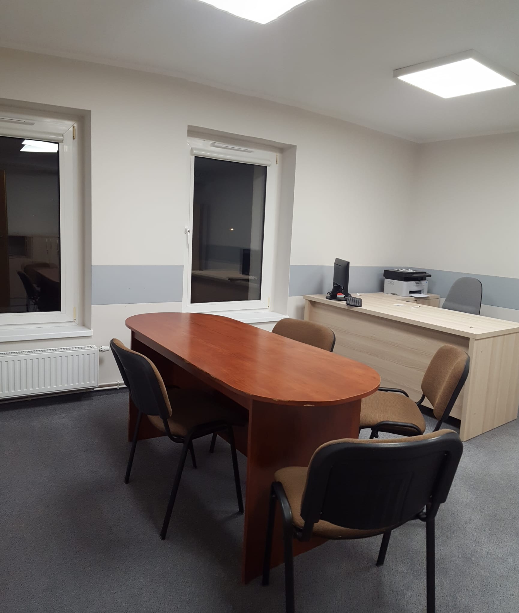 Nowoczesne, jasne biuro po remoncie z owalnym stołem konferencyjnym z brązowego drewna, krzesłami z czarnymi nogami i beżową tapicerką, biurkiem z komputerem i drukarką oraz szarą wykładziną...