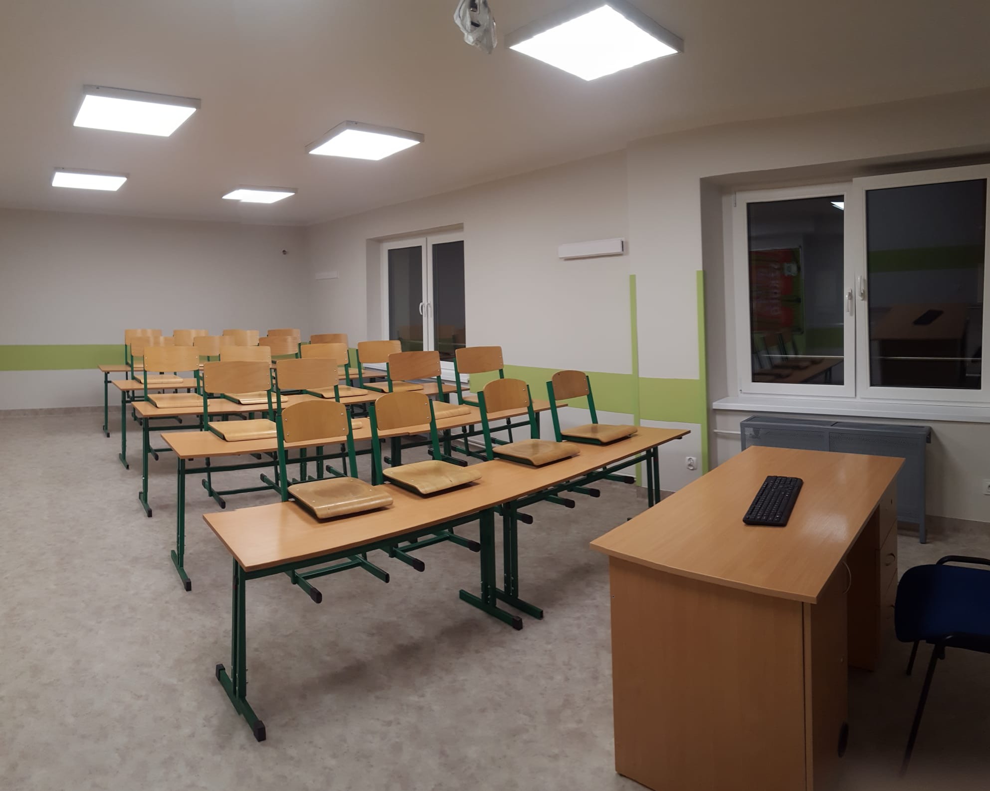Jasna sala lekcyjna z rzędami pustych ławek i krzeseł, biurko nauczycielskie z klawiaturą, równomierne oświetlenie sufitowe.