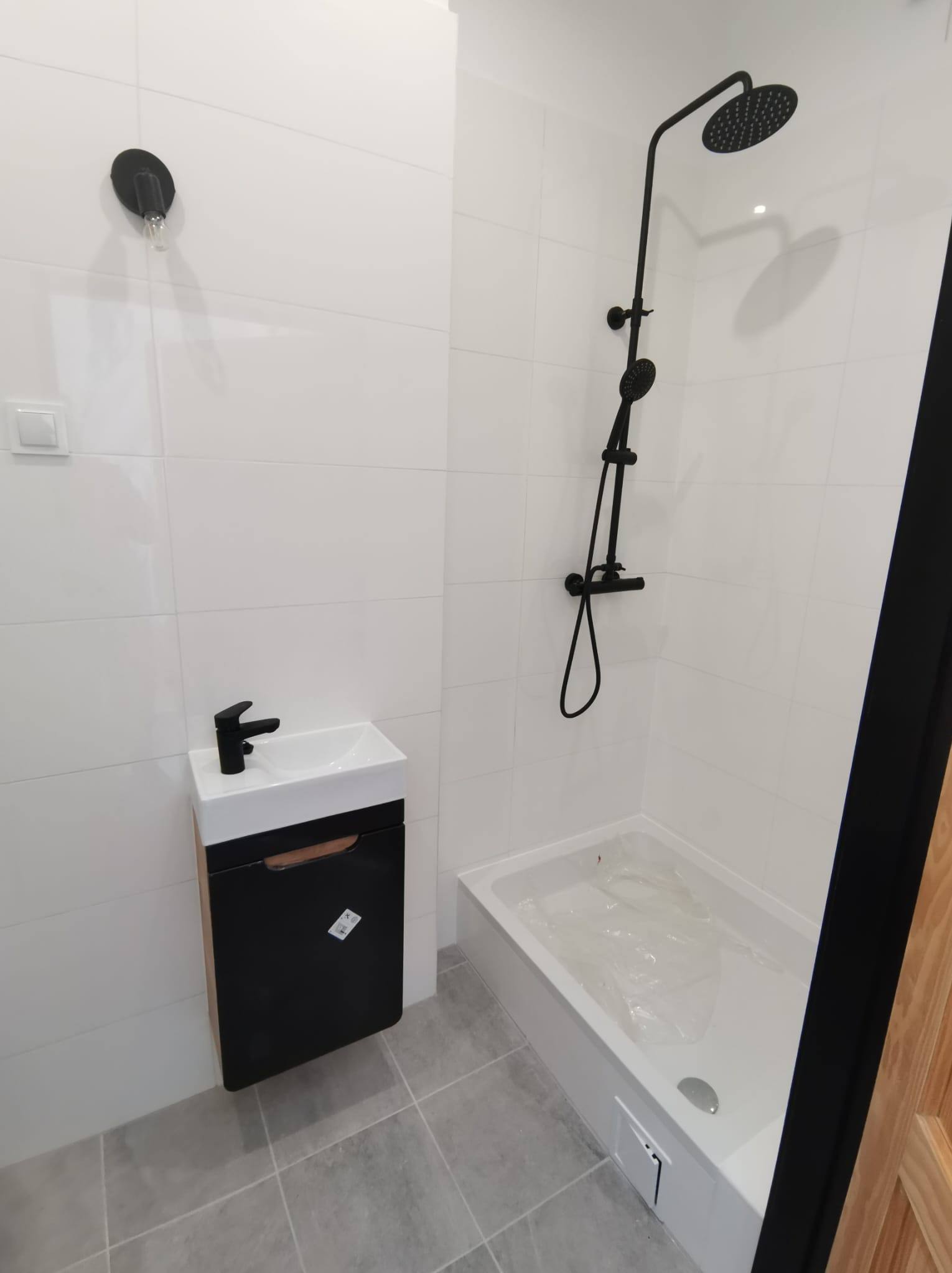 Nowoczesna łazienka z białą armaturą i czarnymi akcentami: umywalka nablatowa z szafką, kabina prysznicowa z deszczownicą, szare płytki podłogowe, minimalistyczny design.