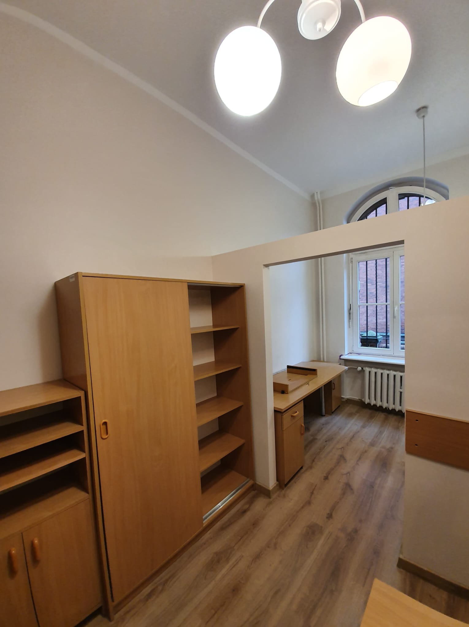 Jasne wnętrze pokoju z drewnianą podłogą, biurkiem przy oknie z kratami, szafami z przesuwnymi drzwiami i oświetleniem sufitowym.