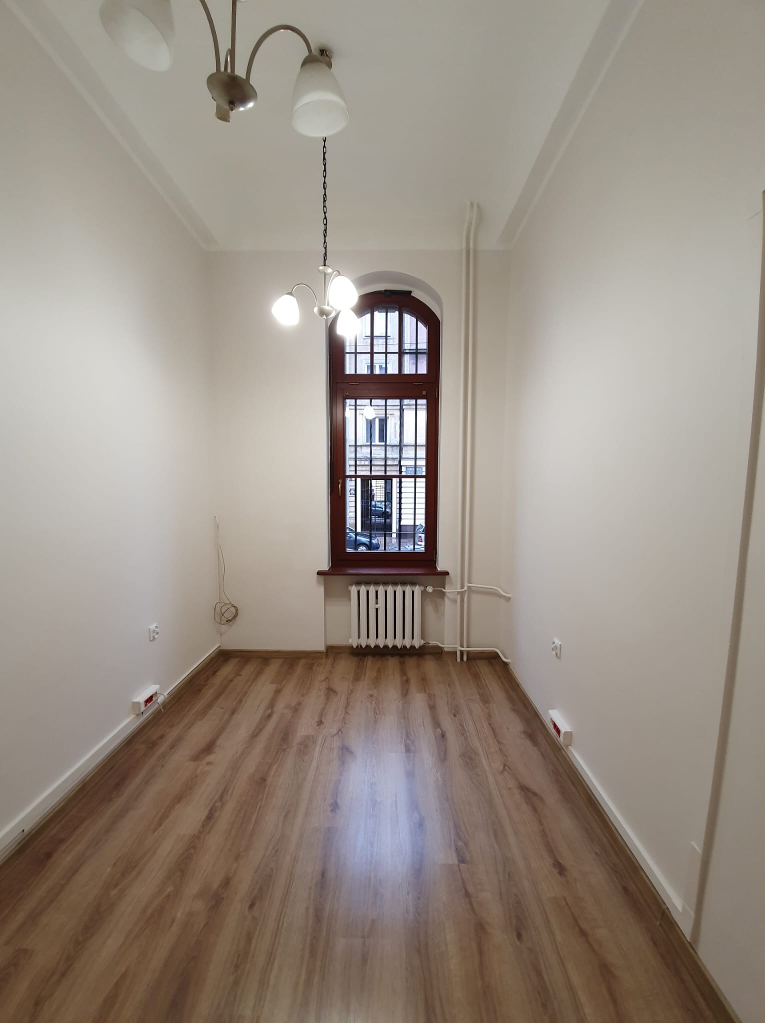 Puste, odnowione wnętrze z drewnianą podłogą, białymi ścianami, grzejnikiem pod oknem w brązowej ramie i żyrandolem w stylu retro.