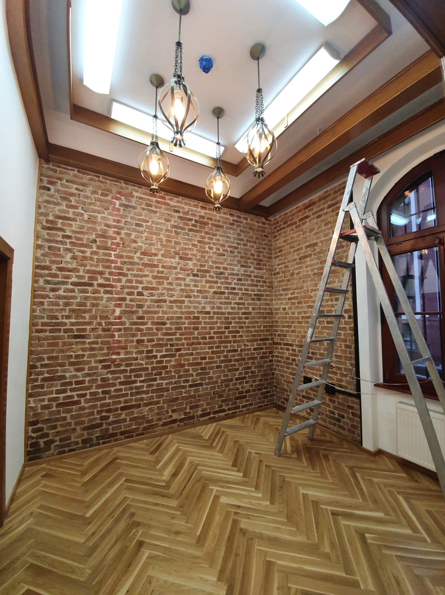 Pokój po remoncie z ceglaną ścianą, parkietem w jodełkę, dekoracyjnymi lampami i rozstawioną drabiną aluminiową.