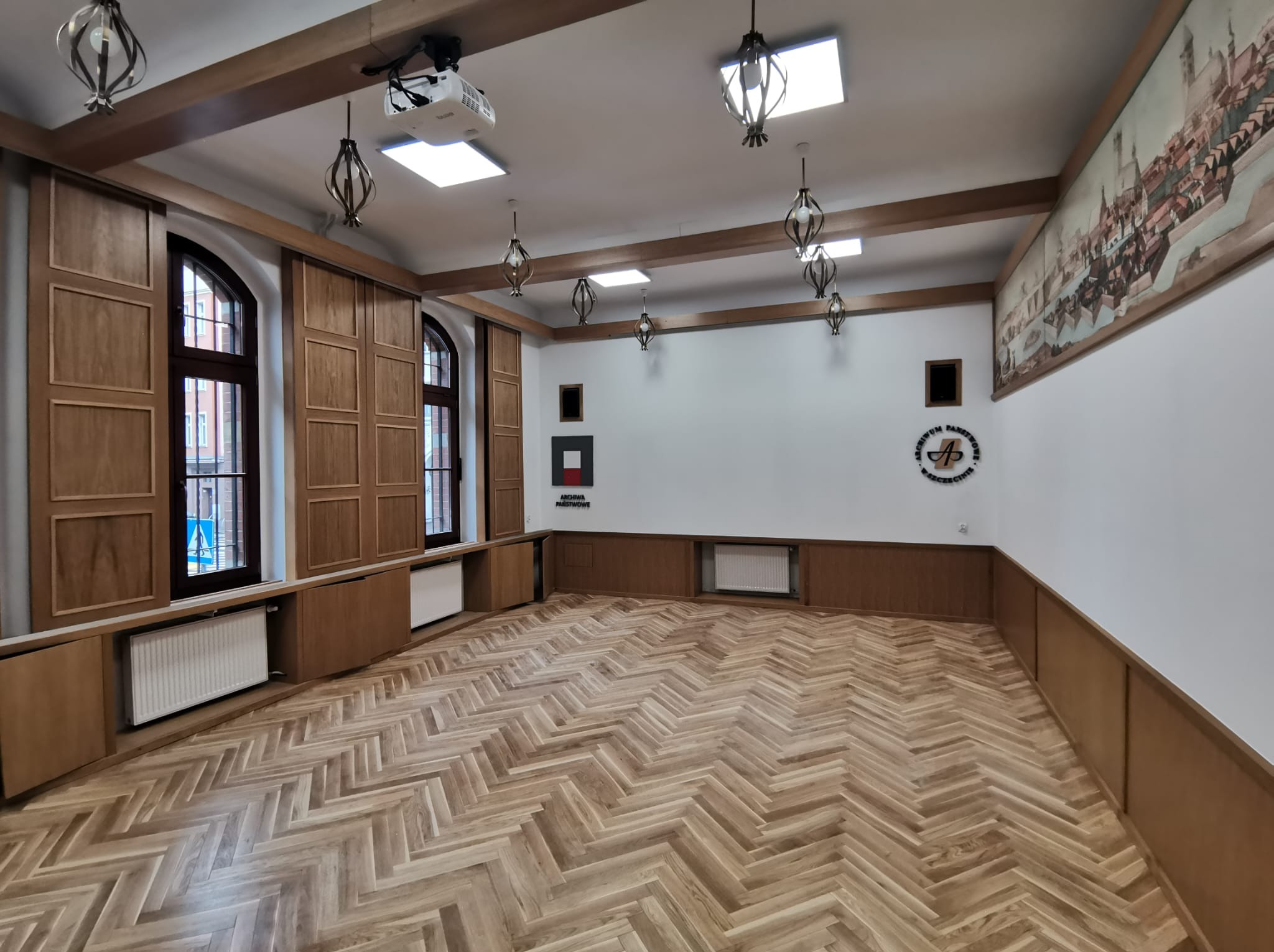 Sala z parkietem ułożonym w jodełkę, drewnianą boazerią, oświetleniem w stylu retro i rzutnikiem pod sufitem. Na ścianie wisi malowidło przedstawiające panoramę miasta.