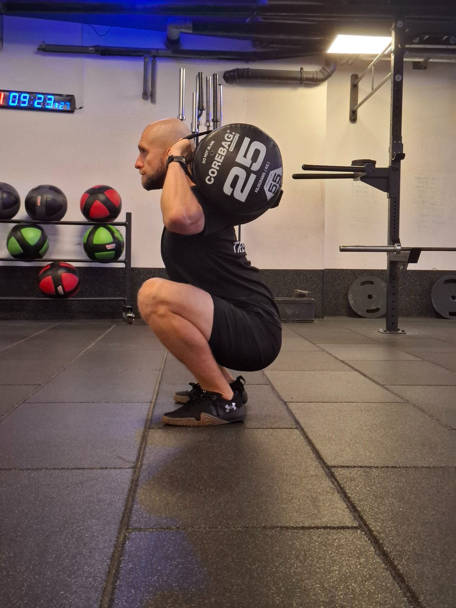Mężczyzna wykonuje przysiad z obciążeniem Corebag 25 kg w siłowni, widok z boku, na ścianie w tle zegar cyfrowy i sprzęt do ćwiczeń