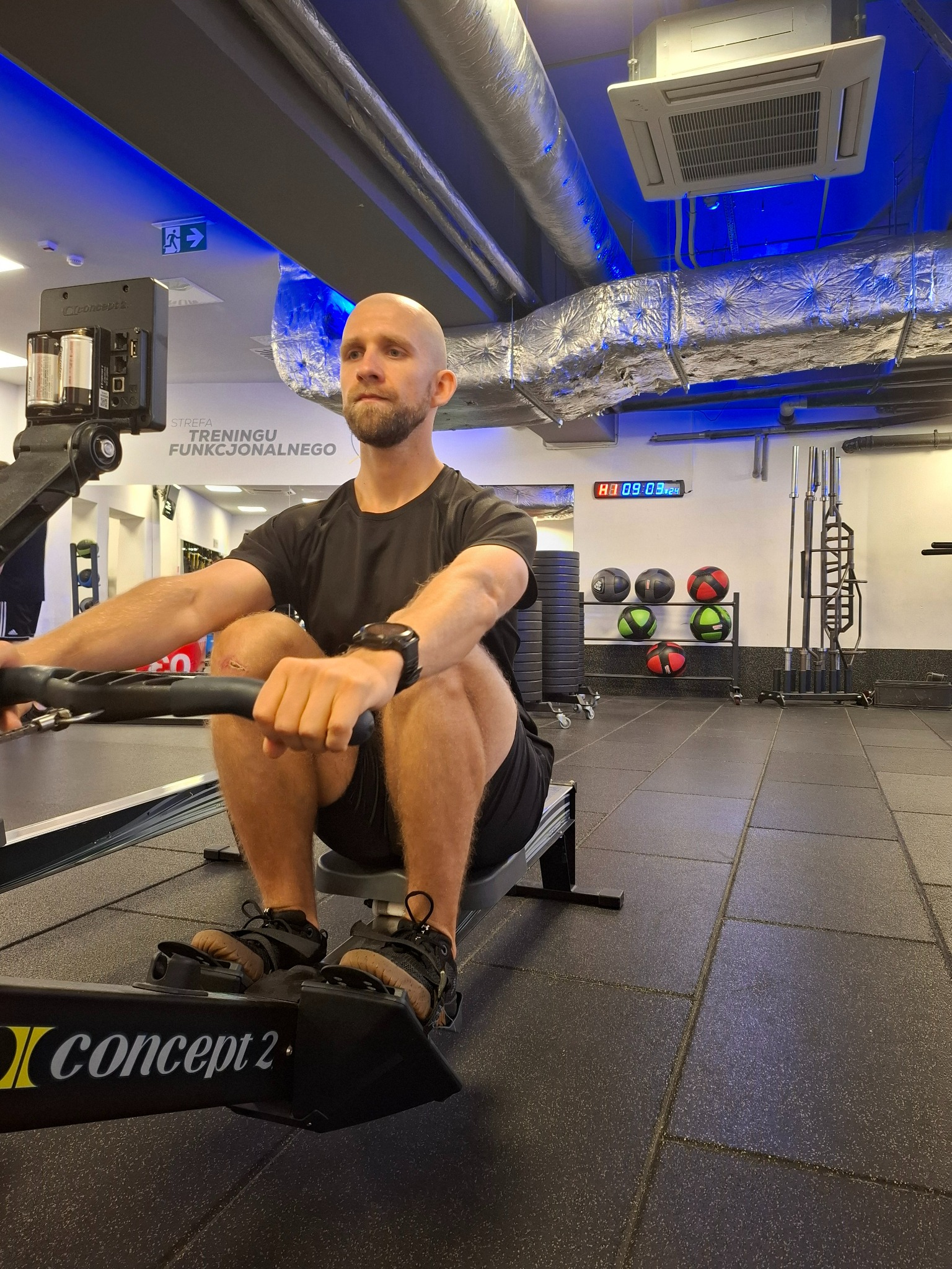 Mężczyzna na ergometrze wioślarskim Concept2 w siłowni z widocznymi piłkami lekarskimi w tle i zegarem cyfrowym na ścianie.