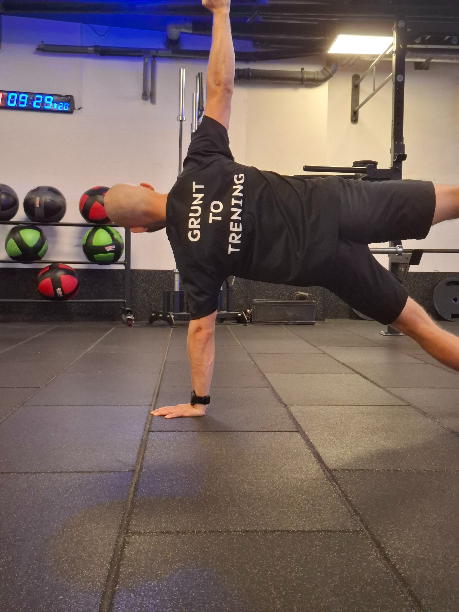 Mężczyzna w czarnej koszulce z napisem 'Grunt to Trening' demonstruje boczną deskę w siłowni na tle piłek lekarskich i sprzętu do ćwiczeń siłowych.