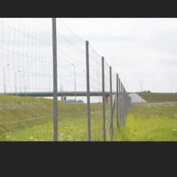 MaxElite - Długa, metalowa siatka ogrodzeniowa z równomiernie rozmieszczonymi słupkami, biegnąca wzdłuż drogi i zielonego terenu, z wiaduktem widocznym w oddali.