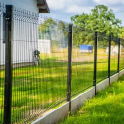 Długi, czarny, metalowy płot panelowy z pionowymi prętami, zamontowany na betonowej podmurówce, biegnący wzdłuż trawnika, z widocznym w tle rowerem opartym o budynek.