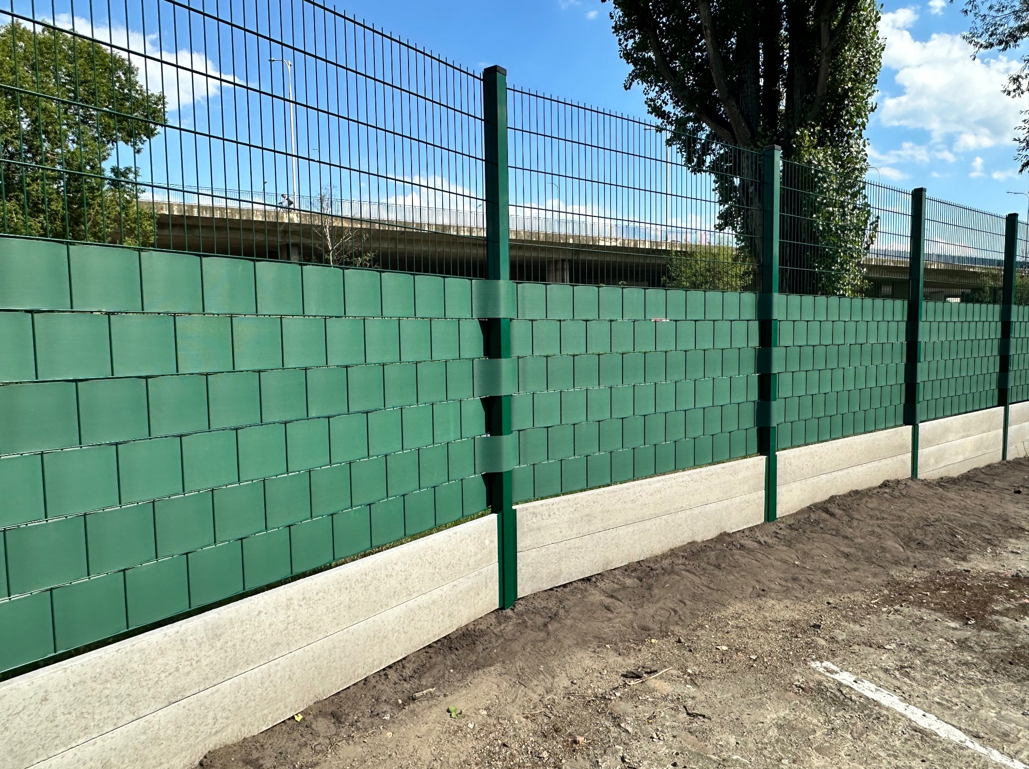 Nowoczesne ogrodzenie panelowe z wypełnieniem w kolorze zielonym, osadzone na betonowej podmurówce, widoczne słupki i stalowa siatka, w tle drzewa i wiadukt.