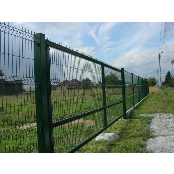 Długi, zielony płot panelowy z siatki, zamontowany na betonowych fundamentach, oddzielający łąkę od drogi żwirowej, z domem w tle i pochmurnym niebem.