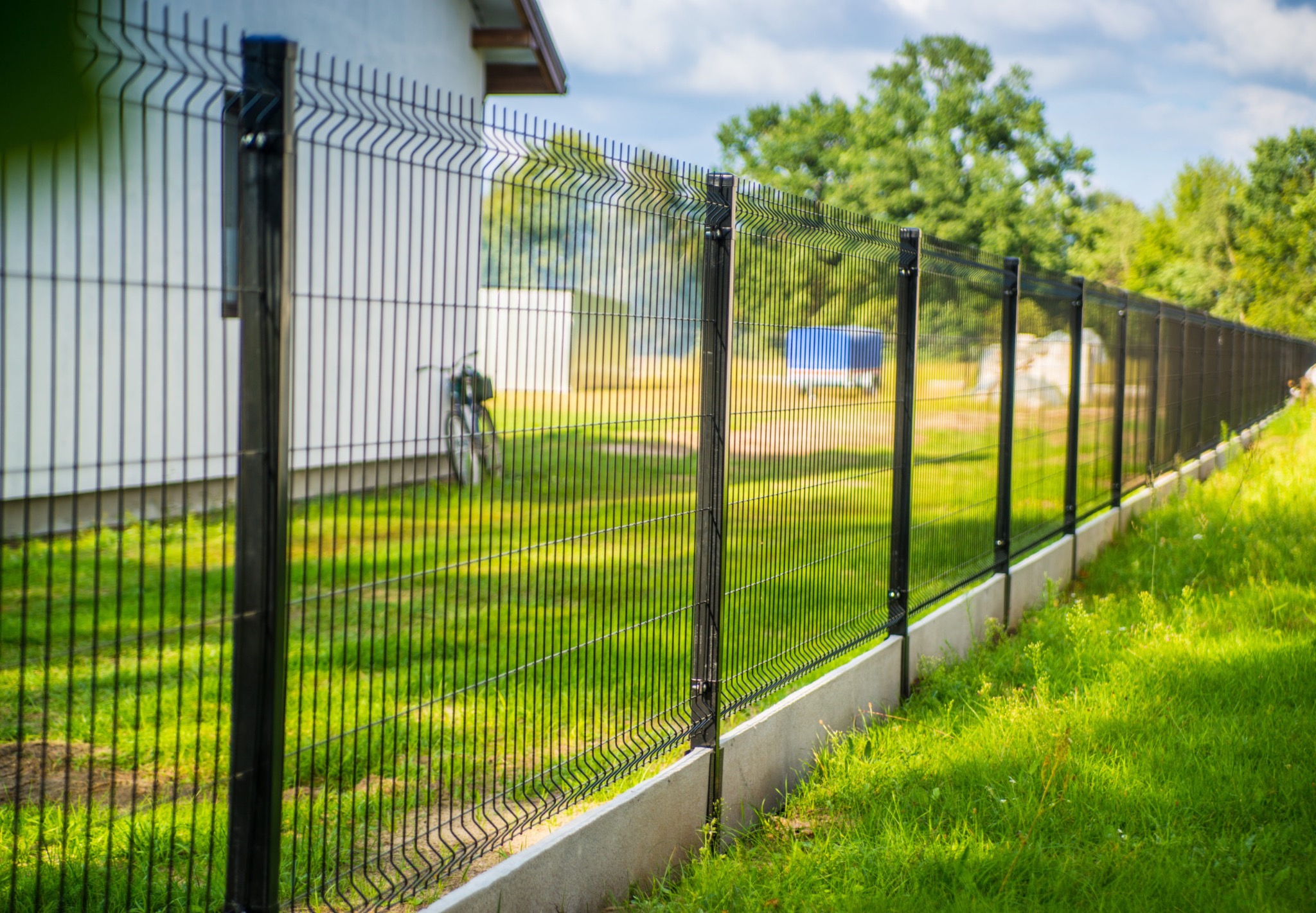 Długi, czarny, metalowy płot panelowy z pionowymi prętami, zamontowany na betonowej podmurówce, biegnący wzdłuż trawnika, z widocznym w tle rowerem opartym o budynek.