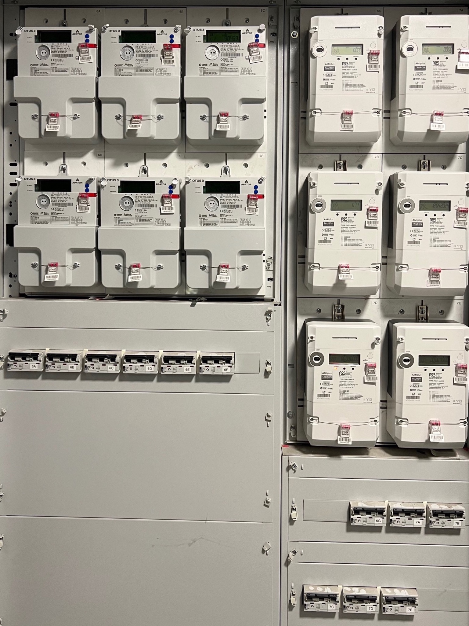 Szara tablica rozdzielcza z rzędami liczników energii elektrycznej różnych producentów i wyłączników oznaczonych numerami, widok z bliska.