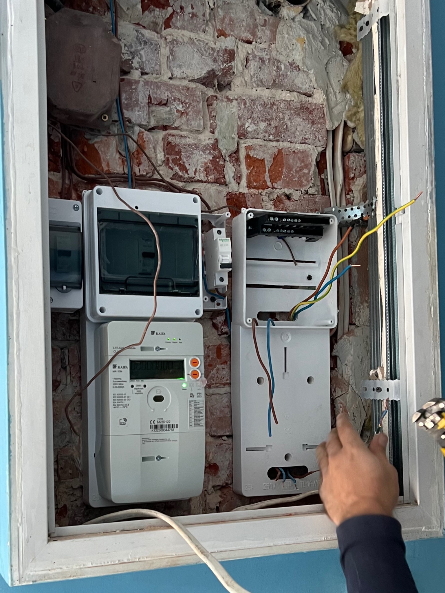 Montaż skrzynki elektrycznej z licznikiem na ścianie z cegły, widoczne okablowanie i ręka elektryka przy pracy. Biała rama okienna dookoła.