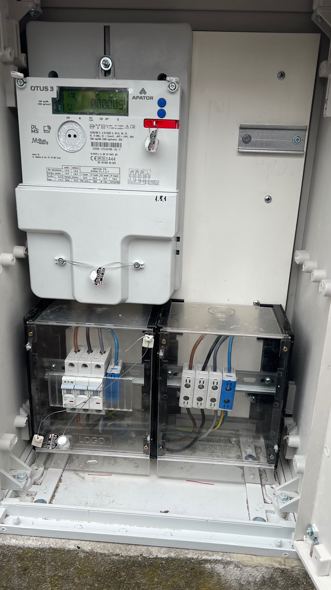 Szczegółowe ujęcie skrzynki elektrycznej z licznikiem Apator QTUS 3, bezpiecznikami EATON i przewodami. Widoczne napisy 'LOGO' i 'WIŚNIEWSKI' na osłonach.