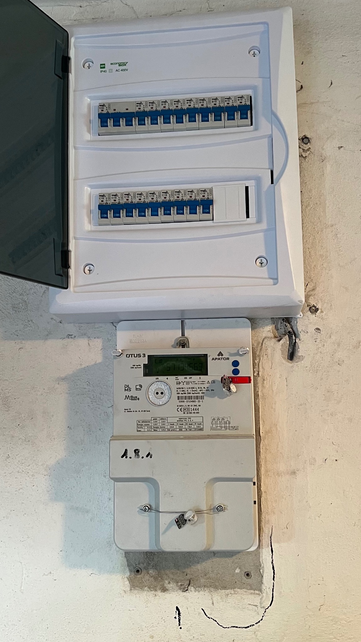 Zamknięta skrzynka bezpiecznikowa i licznik energii elektrycznej na ścianie. Widoczne zabezpieczenia i oznaczenia na urządzeniach.