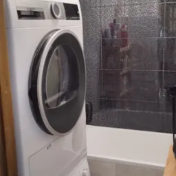 Ustawiona pionowo pralka i suszarka Bosch w łazience z mozaiką na ścianie, widoczna wanna i akcesoria łazienkowe.