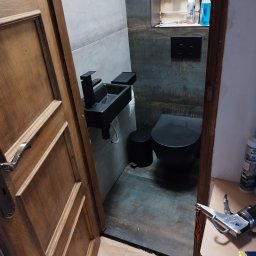 Usługi Monterskie Mikołaj Śledź - Nowoczesna, czarna armatura łazienkowa: umywalka i toaleta, na tle szaro-grafitowych płytek, widoczne narzędzia glazurnicze i otwarte drewniane drzwi.