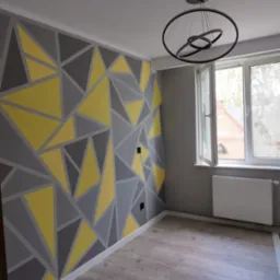 Pokój po remoncie z szaro-żółtą geometryczną dekoracją malarską na ścianie, nowoczesna lampa wisząca i otwarte okno z widokiem na ceglany budynek.