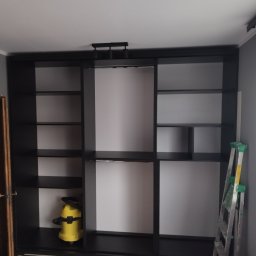 Usługi Monterskie Mikołaj Śledź - Czarna szafa wnękowa z przesuwnymi drzwiami, częściowo pusta, z żółtym odkurzaczem przemysłowym w jednej z półek i aluminiową drabiną opartą o ścianę po prawej stronie.