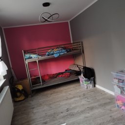 Usługi Monterskie Mikołaj Śledź - Pokój dziecięcy z metalowym łóżkiem piętrowym na tle ściany pomalowanej na różowo i szaro, widoczne panele podłogowe i nowoczesna lampa sufitowa.