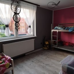 Usługi Monterskie Mikołaj Śledź - Pokój dziecięcy z łóżkiem piętrowym, ścianą w kolorze fuksji i szarą, drewnianą podłogą. Na oknie białe zasłony z czarnymi ozdobami i łapacz snów.