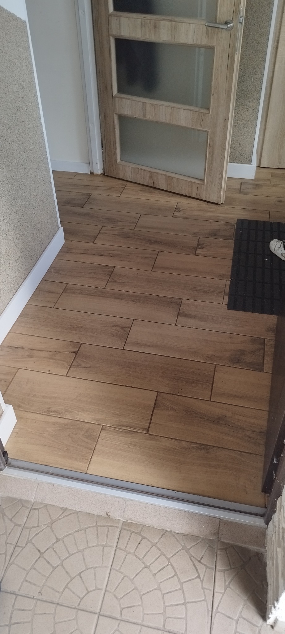 Ułożona podłoga z płytek imitujących drewno, widoczna przy progu drzwi z matową szybą i jasnymi ościeżnicami, z fragmentem dywanika przy drzwiach.