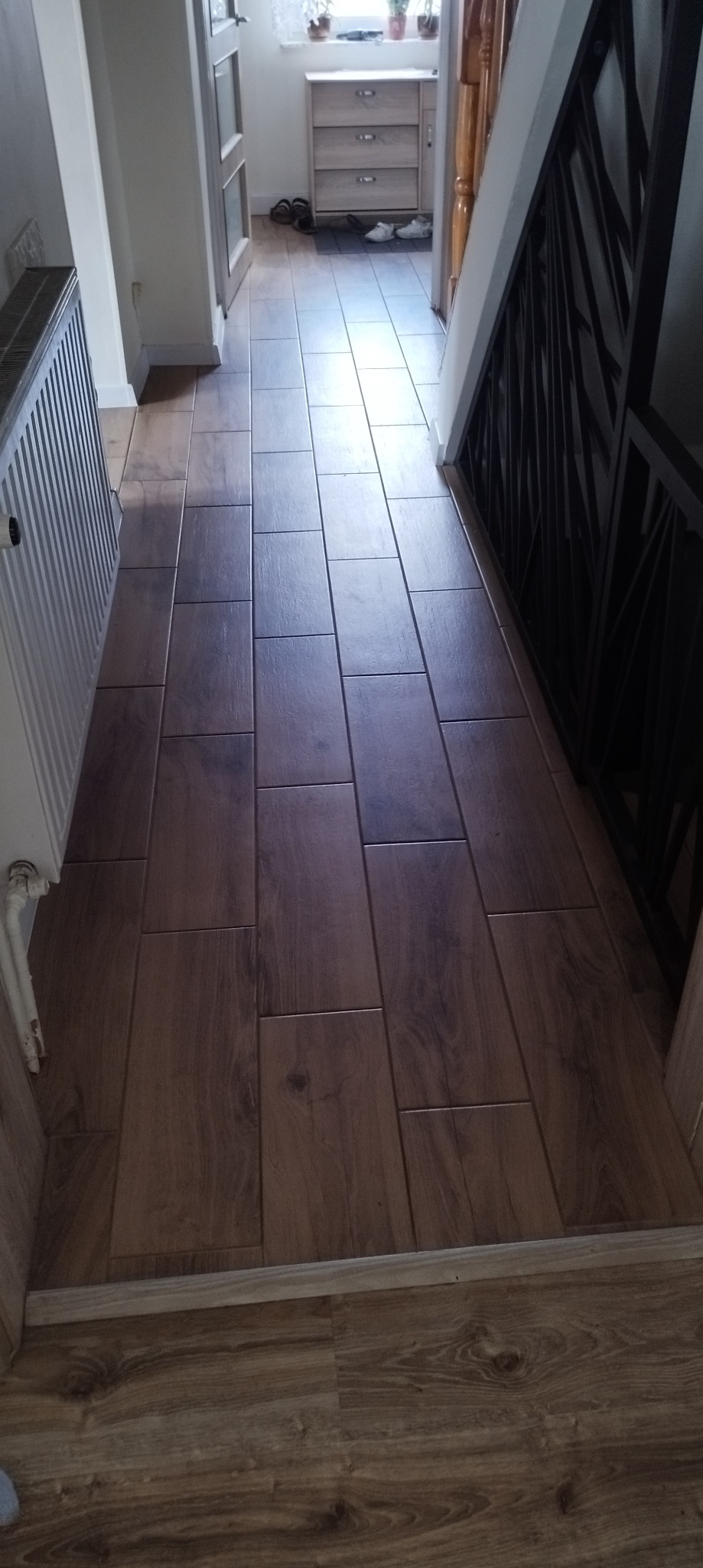 Korytarz z nową podłogą wyłożoną podłużnymi płytkami imitującymi drewno, schody z czarną metalową balustradą po prawej, grzejnik po lewej stronie, w tle widoczny fragment szafki i drzwi.
