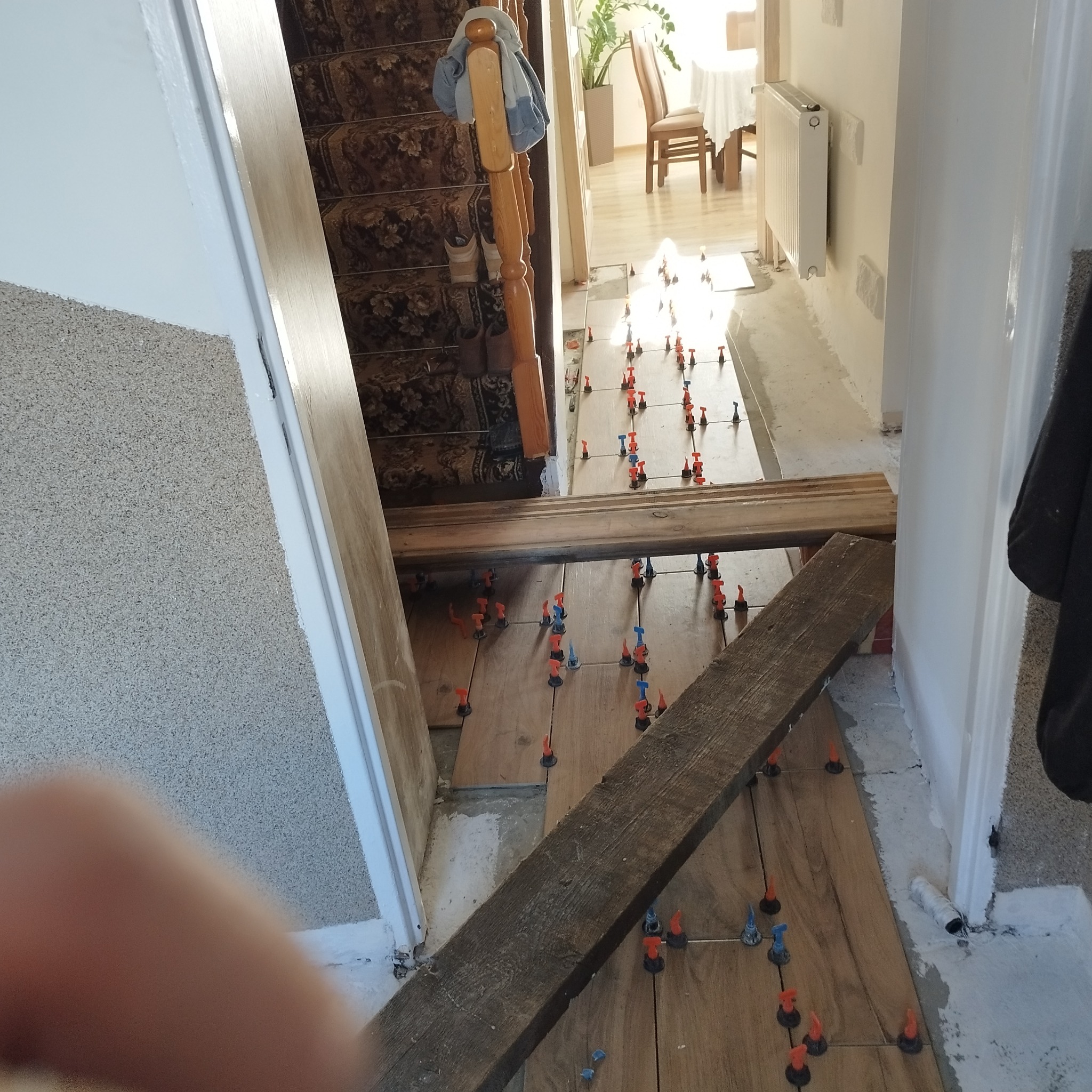 Kładzenie drewnopodobnych płytek w korytarzu, widoczne poziomice i klipsy poziomujące, schody w tle, surowe wykończenie ścian.