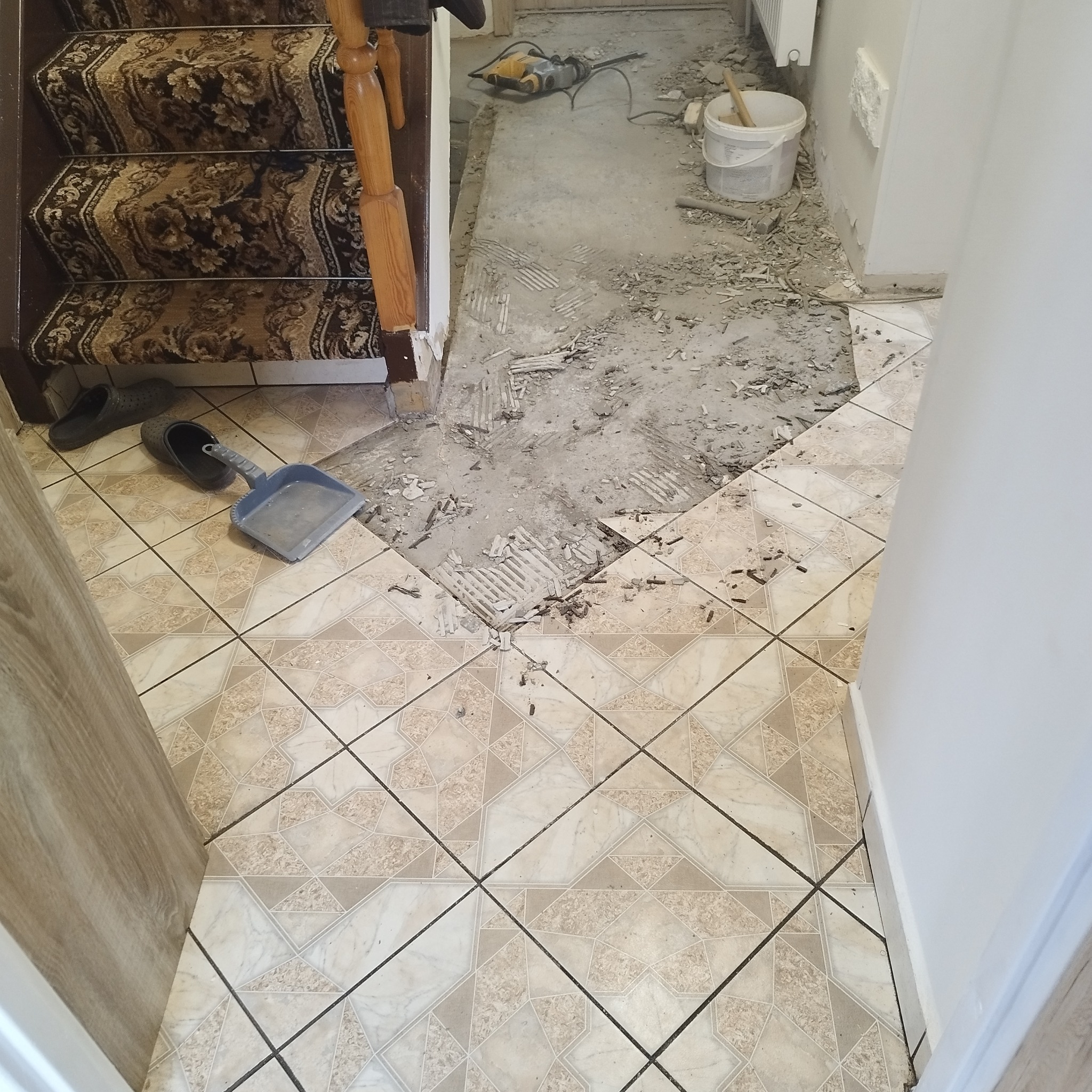Demontaż starej podłogi z płytek przed schodami, widoczne narzędzia, gruz i fragment nowej podłogi z jasnych płytek w geometryczny wzór.