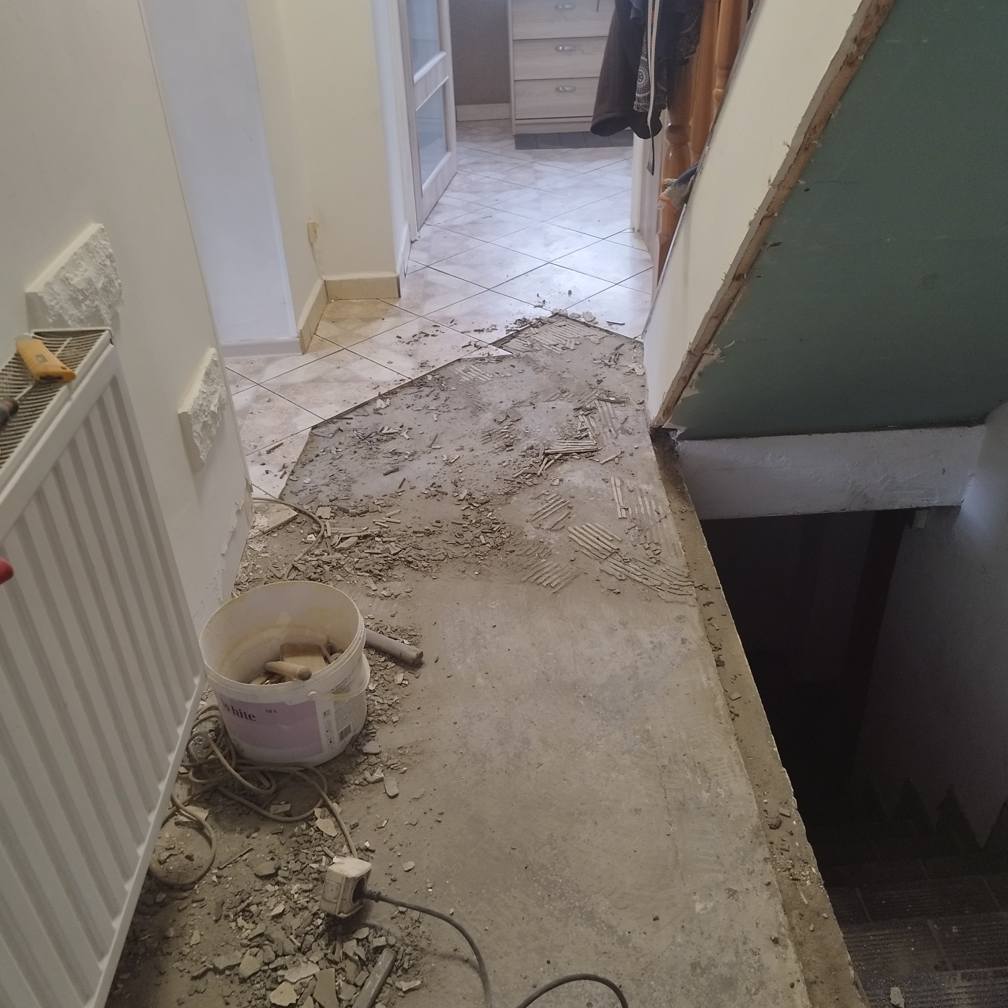Demontaż płytek podłogowych w korytarzu, widoczny fragment schodów, resztki kleju i gruzu na podłodze, białe wiadro z narzędziami obok grzejnika.