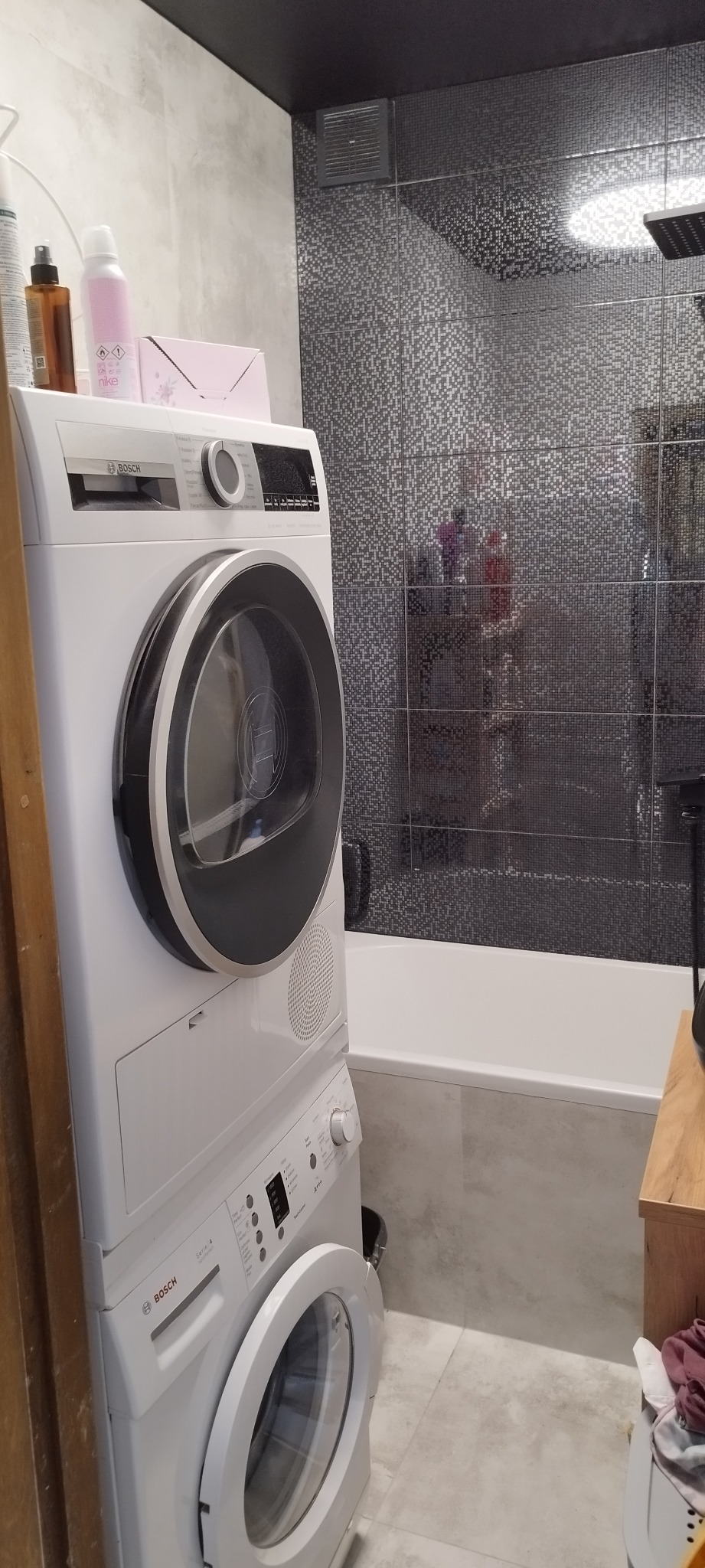 Ustawiona pionowo pralka i suszarka Bosch w łazience z mozaiką na ścianie, widoczna wanna i akcesoria łazienkowe.