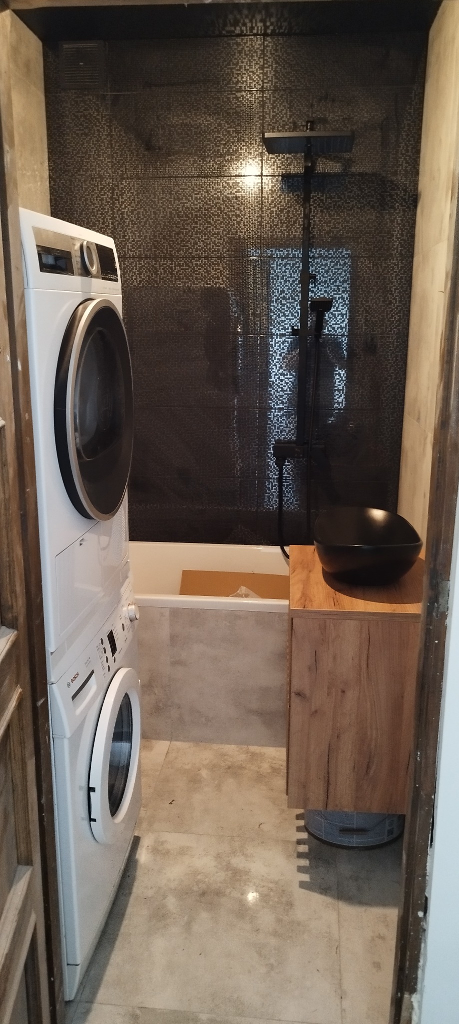 Nowoczesna łazienka z ciemnymi mozaikowymi płytkami, zestawem pralko-suszarki Bosch, czarną umywalką na drewnianej szafce i baterią prysznicową w kolorze czarnym, widok z wejścia.