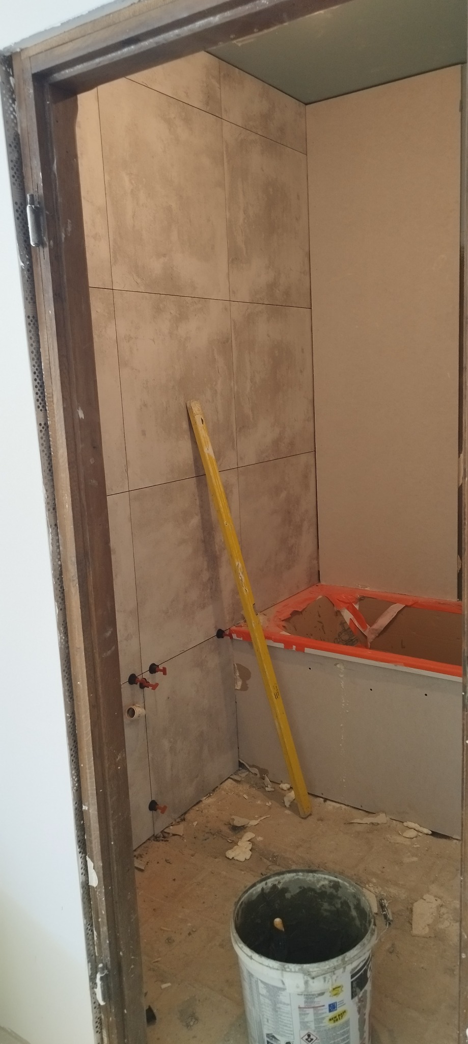 Prace w toku w łazience: częściowo wyłożone płytki na ścianie, poziomica oparta o ścianę, widoczne elementy instalacji wodnej, przygotowana obudowa wanny, wiadro z zaprawą na podłodze.